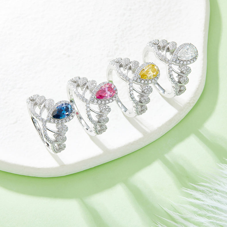 CARISSIE pear moissanite halo ring lineup, diagonal display of blue, pink, yellow and white stones on textured white surface