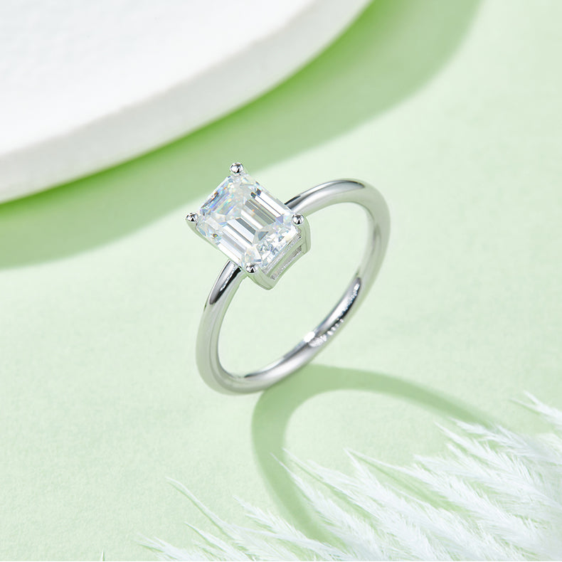 CARISSIE emerald cut moissanite solitaire ring, lifestyle shot on a soft green surface with white feathers adding texture to the scene