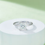 CARISSIE Princess Solitaire Engagement Ring, elevated display on white podium against a pastel green background